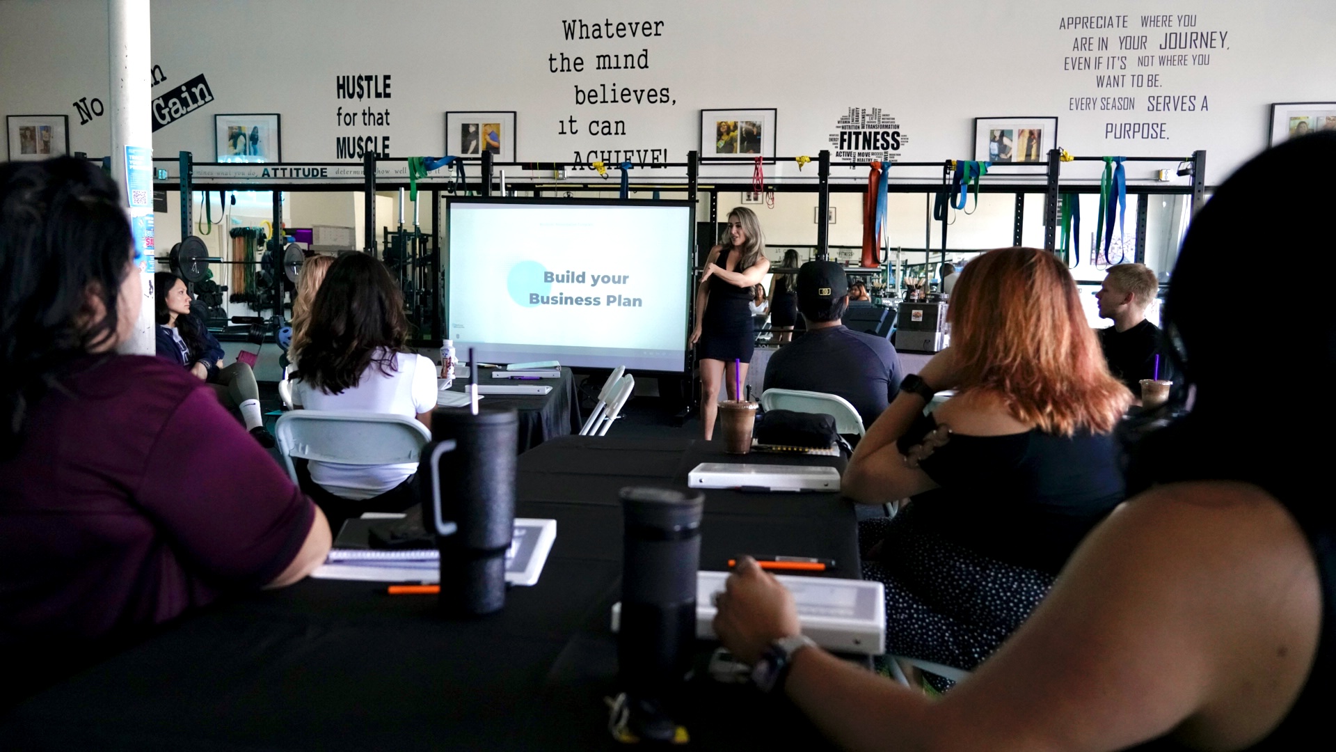 People seated at tables in a classroom or conference room, facing a presenter and a screen, with motivational quotes on the wall.