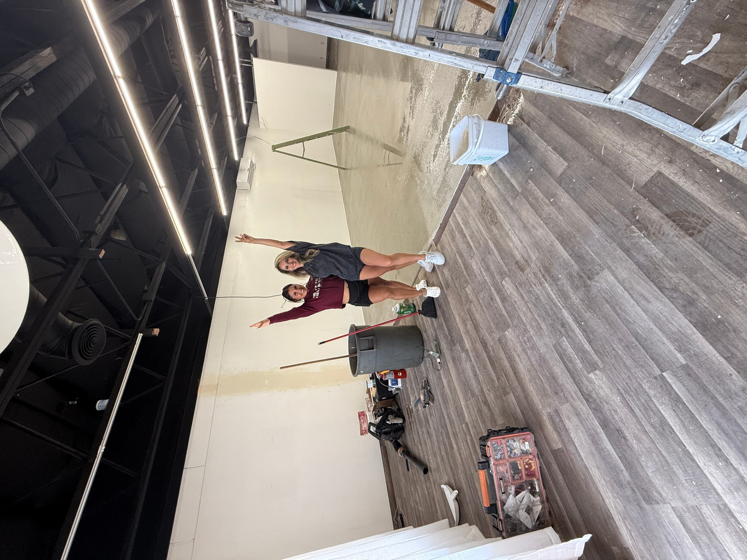 Two people stand on a wooden floor in a large room under construction, with construction materials and tools around.