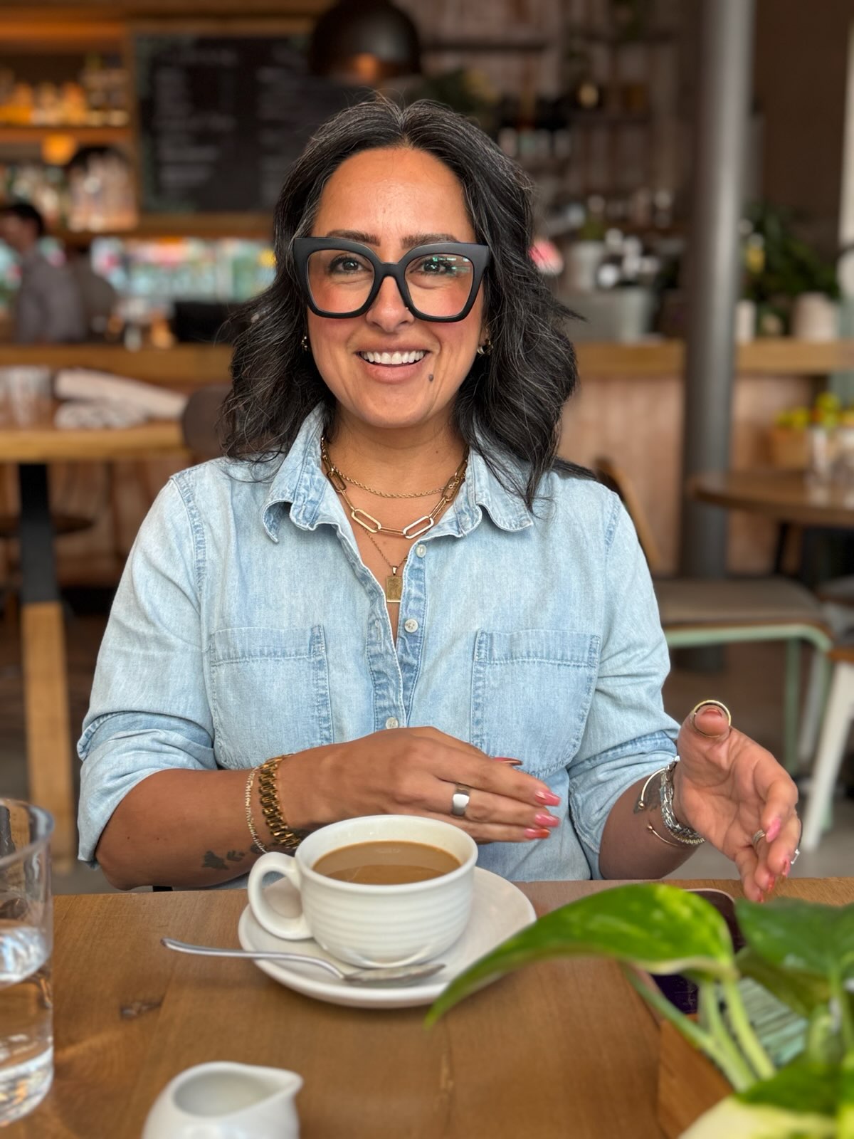 Woman with glasses smiling at a cafe, sitting at a table with a cup of coffee, wearing a light denim shirt.