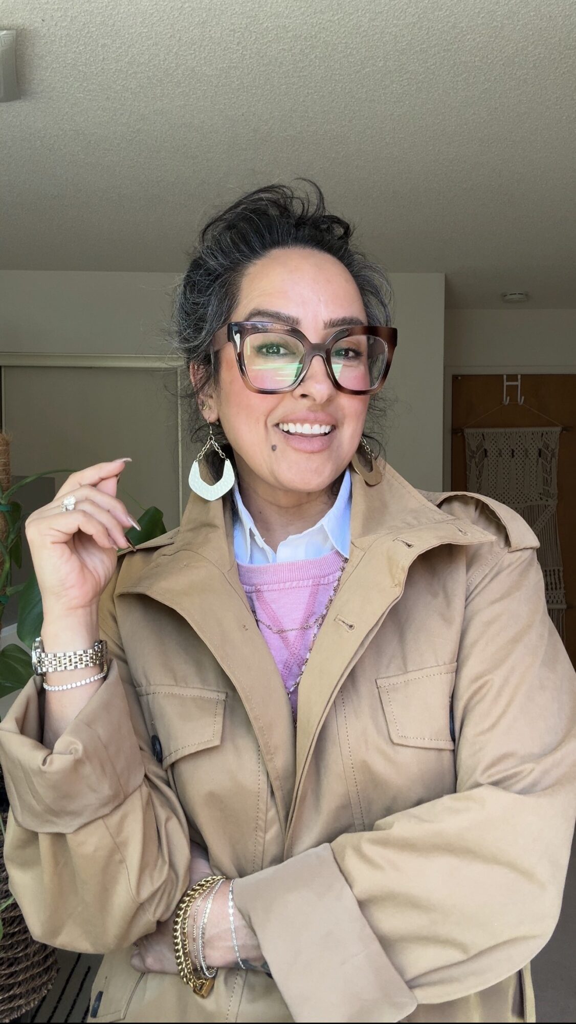 Woman with glasses smiling, wearing a beige jacket and jewelry, indoors with a neutral background.