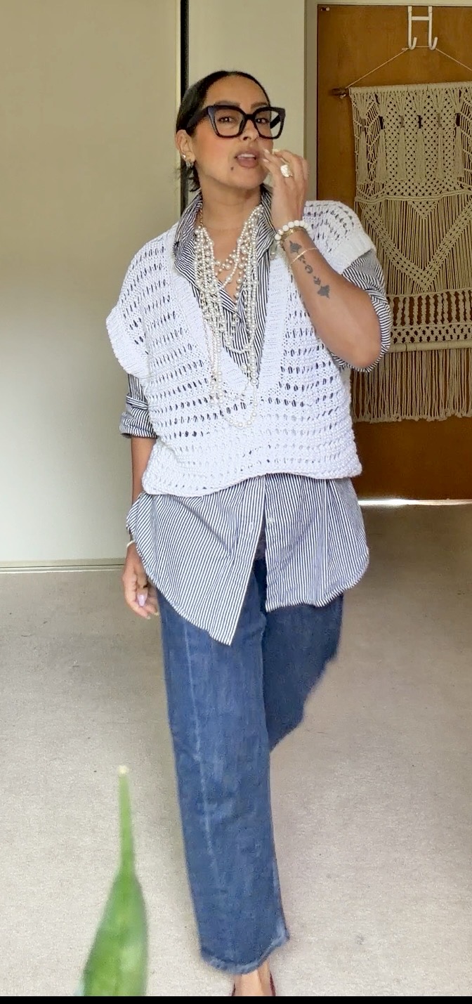Woman wearing glasses, layered clothing, standing indoors, with a decorative wall hanging behind her.