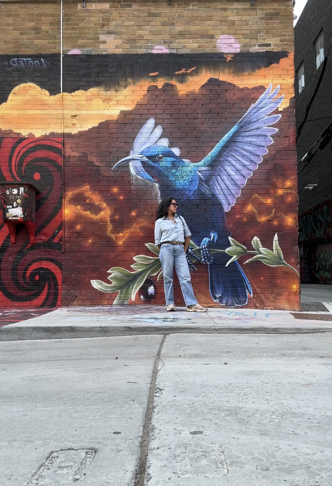 Person standing in front of a colorful mural of a blue hummingbird with outstretched wings, clouds, and abstract designs on a brick wall.