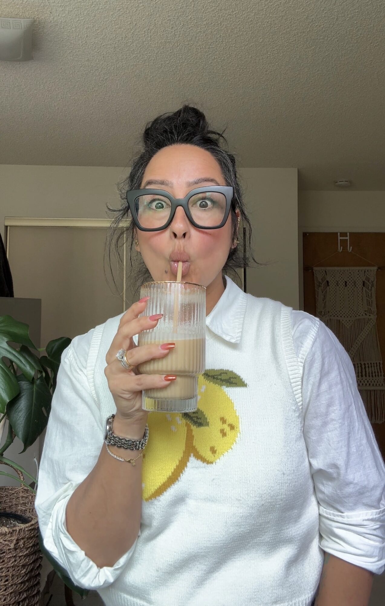 Woman with glasses and dark hair in a bun drinks from a glass with a lemon slice, wearing a white shirt.
