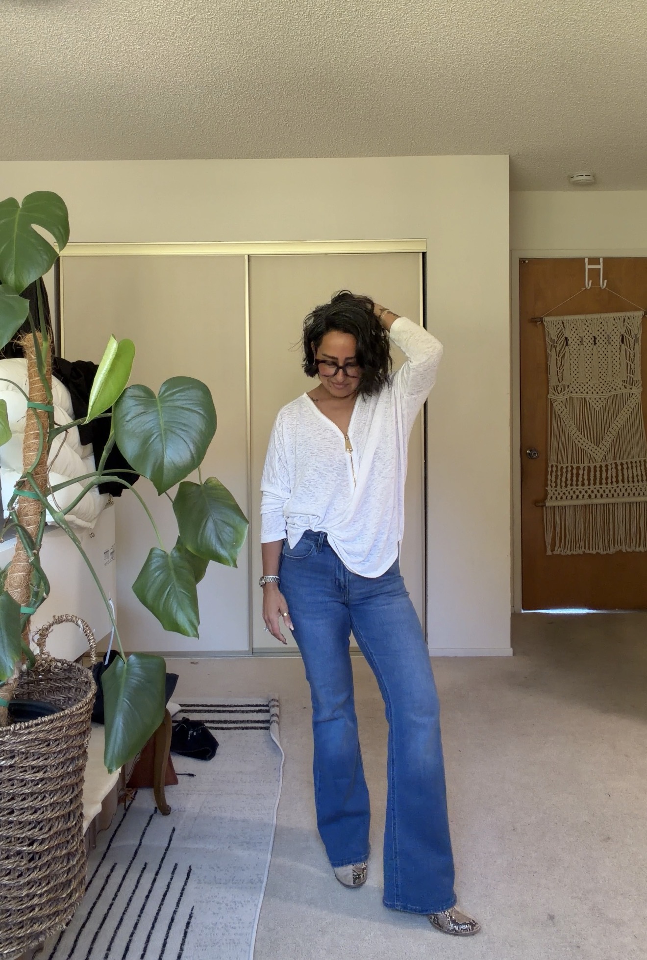 Woman with curly hair and glasses stands in a room, wearing a white blouse and blue jeans, with a large plant nearby.