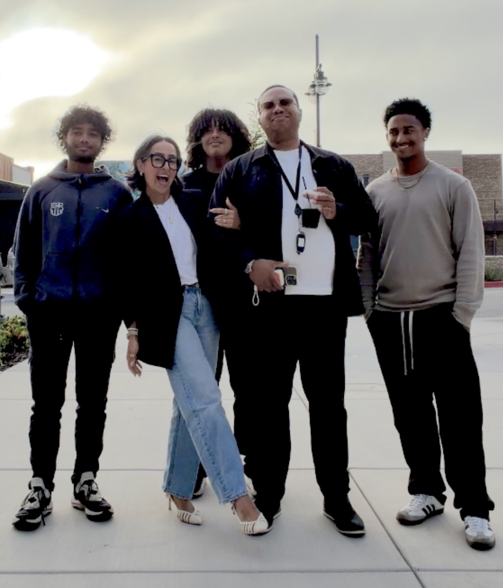 Group of five people standing outdoors, some with arms crossed, with a tower in the background, cloudy sky, and sunlight.
