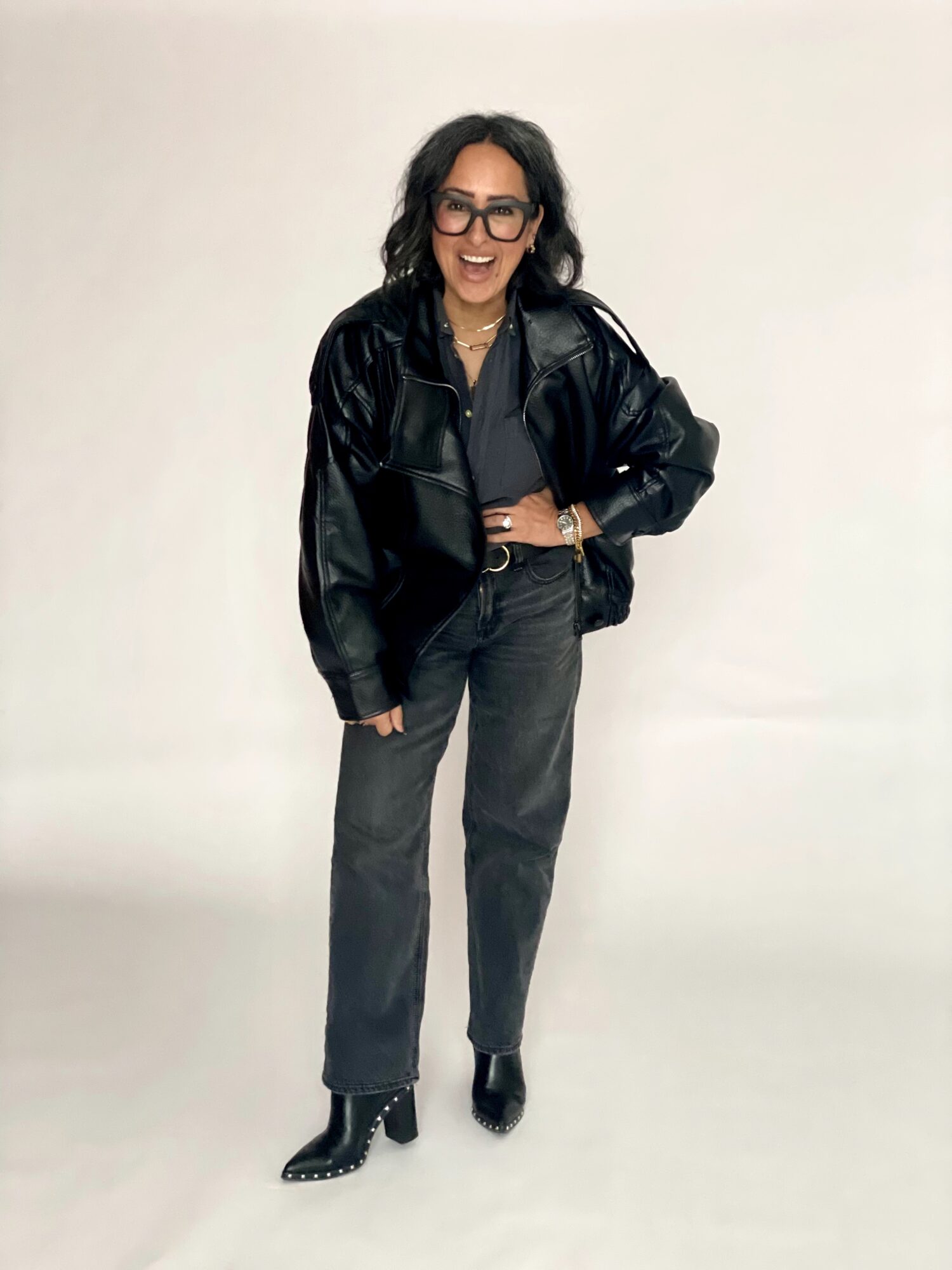 Woman with dark hair and glasses wearing a black jacket, gray pants, and black boots, smiling against a plain background.