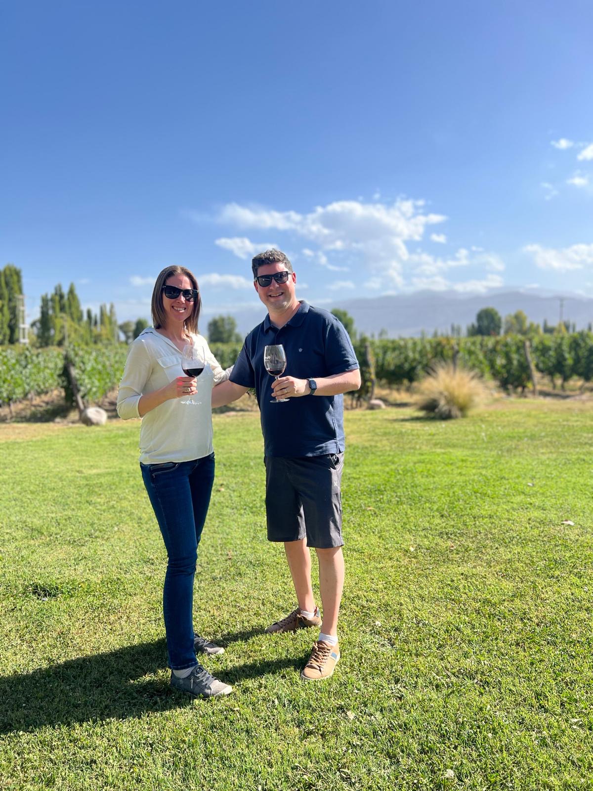 Two people stand on a grassy field holding wine glasses, with trees and a blue sky in the background.