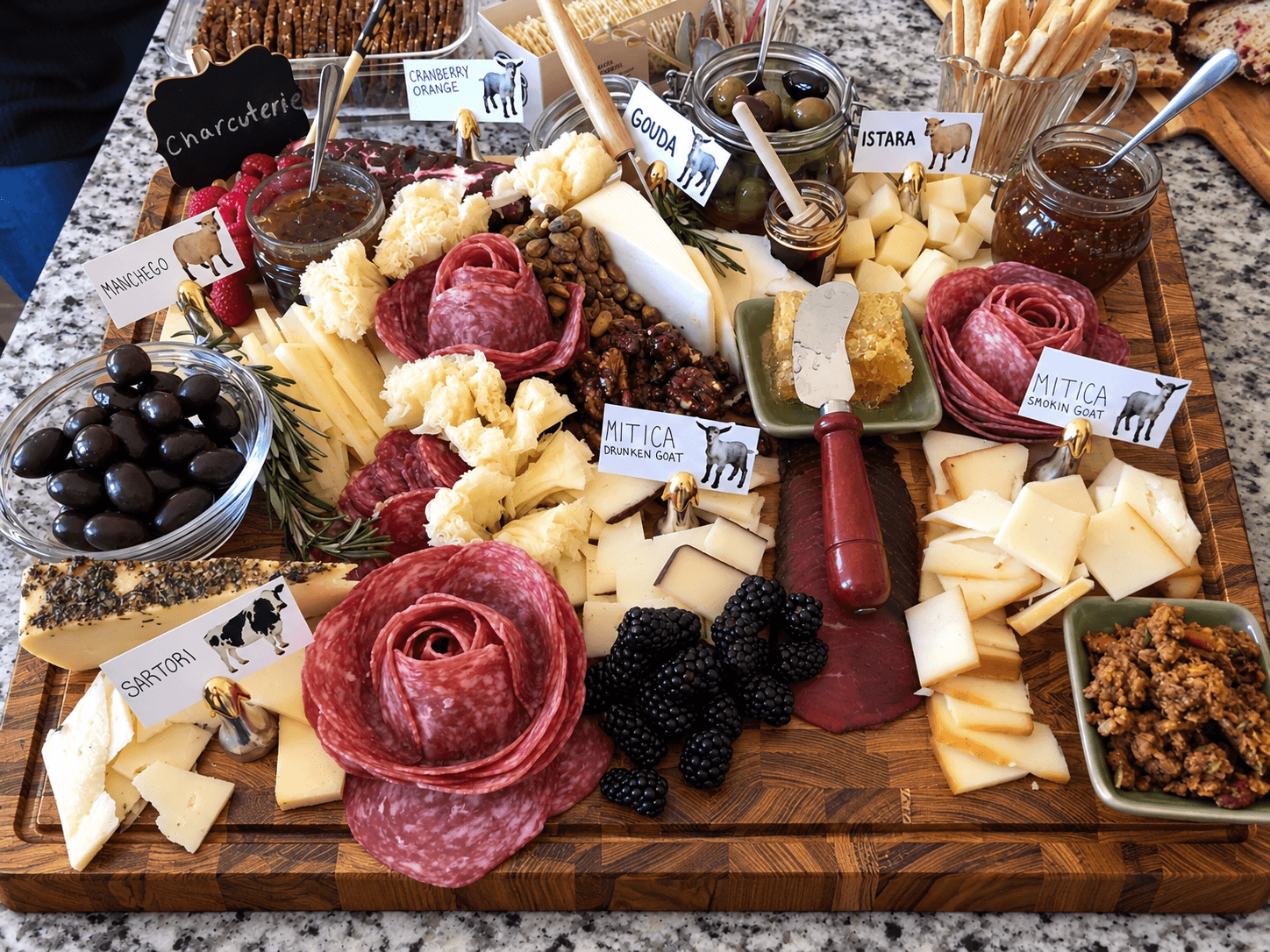 Cheese, meats, grapes, and condiments on a wooden board with small labels, surrounded by breadsticks and jars.