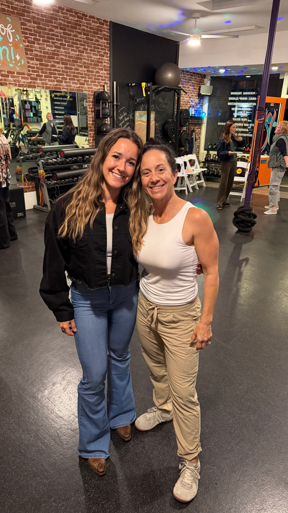 Two women standing together in a gym, smiling, with workout equipment and mirrors in the background.