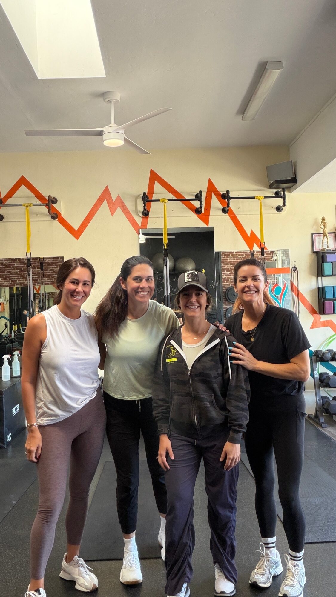 Four women standing together indoors, smiling, with fitness equipment and a red zigzag line on the wall behind them.