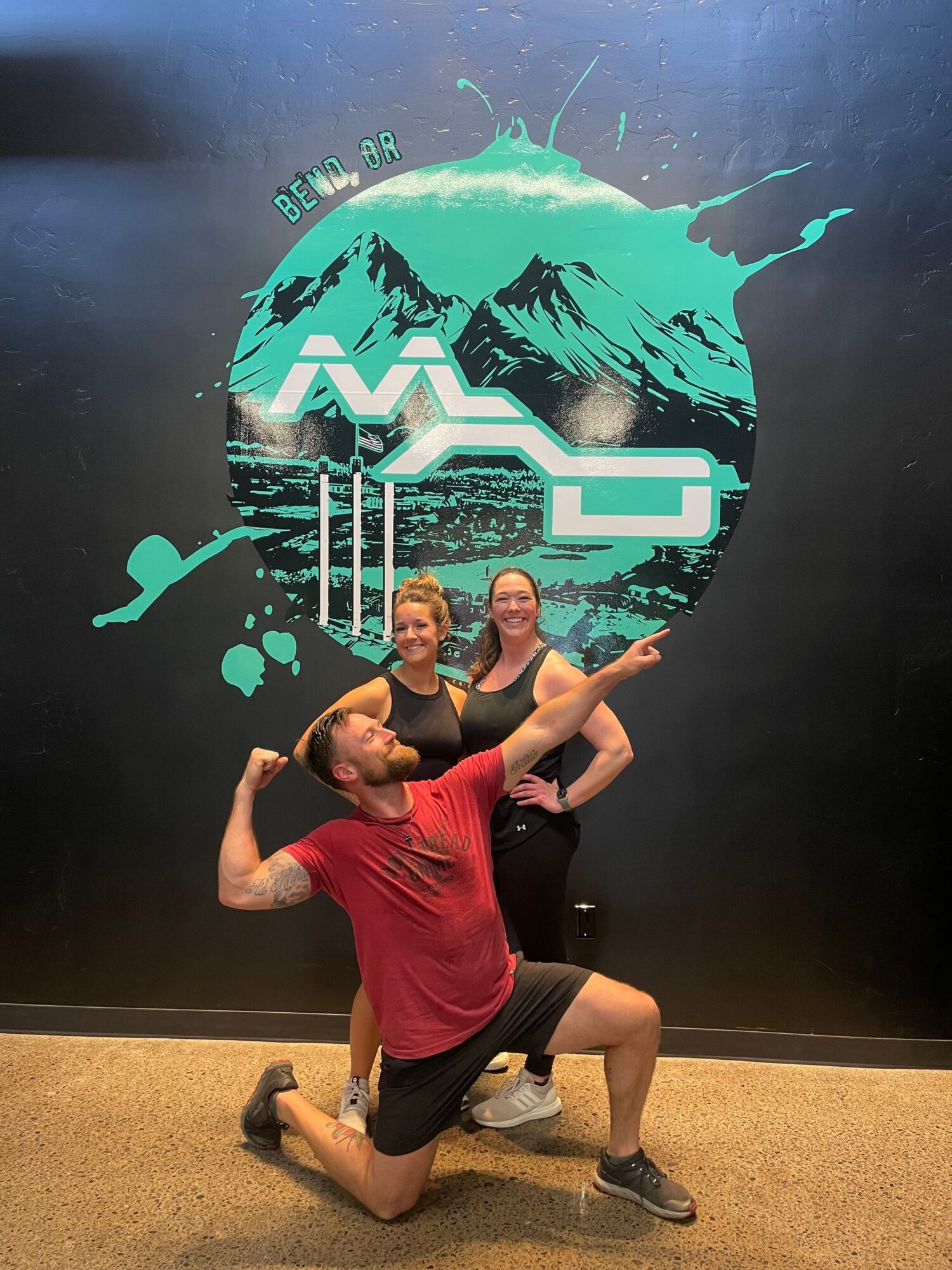Two people pose in front of a graffiti-style mural with a large circular design and abstract elements, one kneeling and one standing.