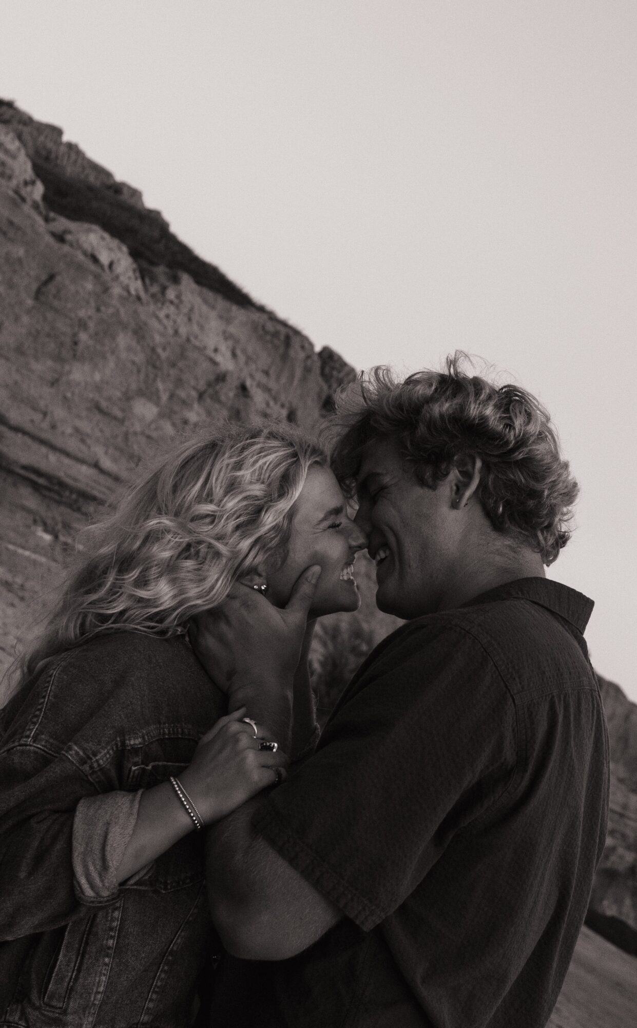 Two people with curly hair face each other closely outdoors, with a rocky hillside in the background.