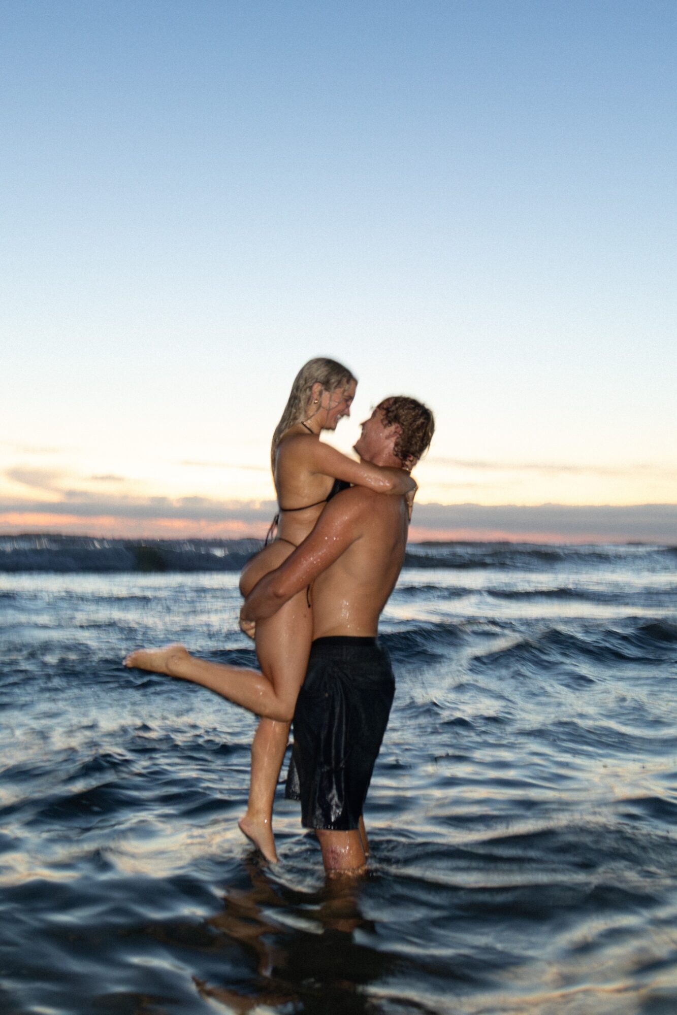 A man lifts a woman in the ocean with waves, both are topless, during sunset sky, water, and horizon visible.