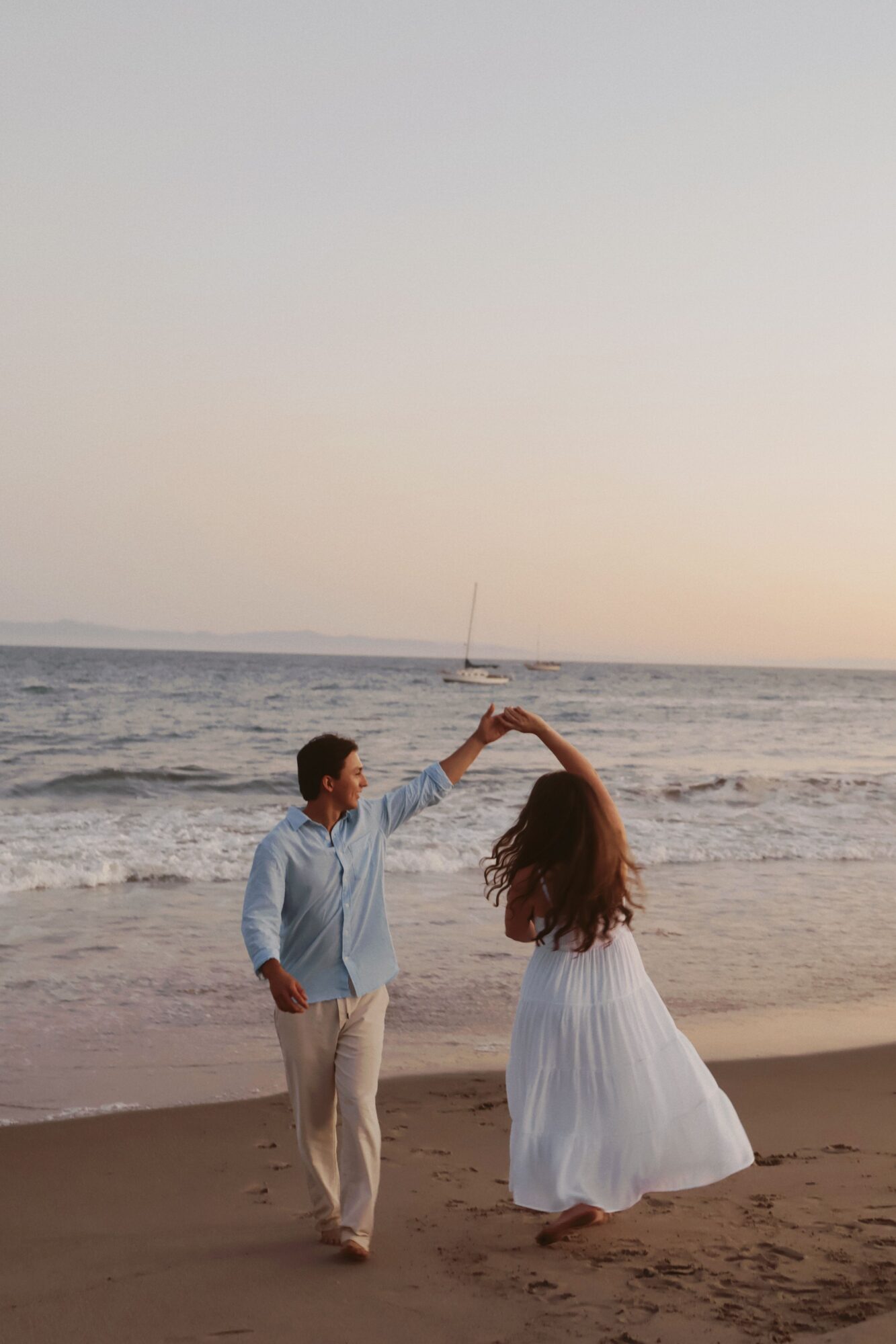 A woman in a white dress and a man in light-colored clothing stand on a beach at sunset, holding hands and looking at the ocean.