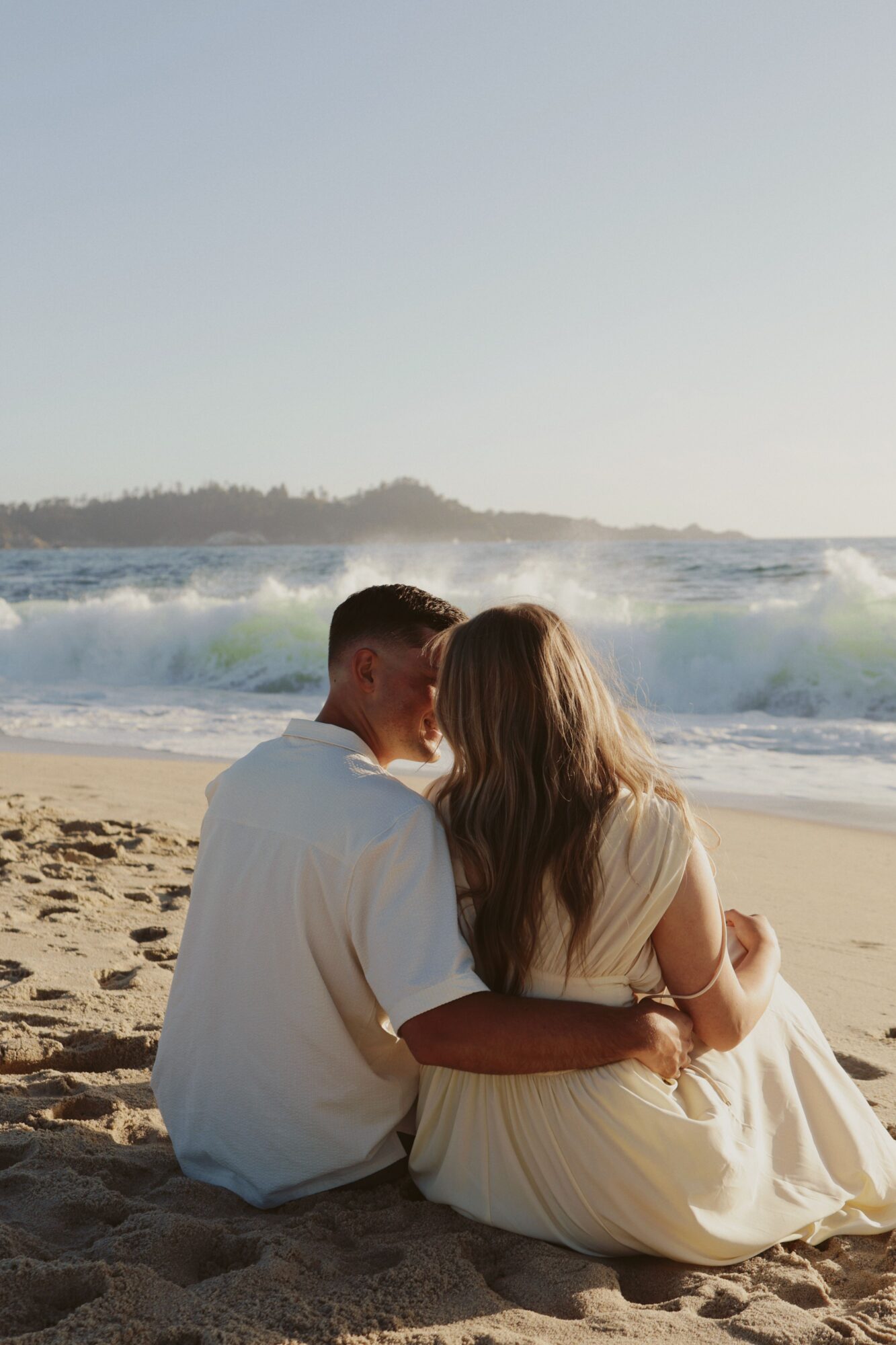 Two people sit on a sandy beach facing the ocean, embracing each other with waves in the background.