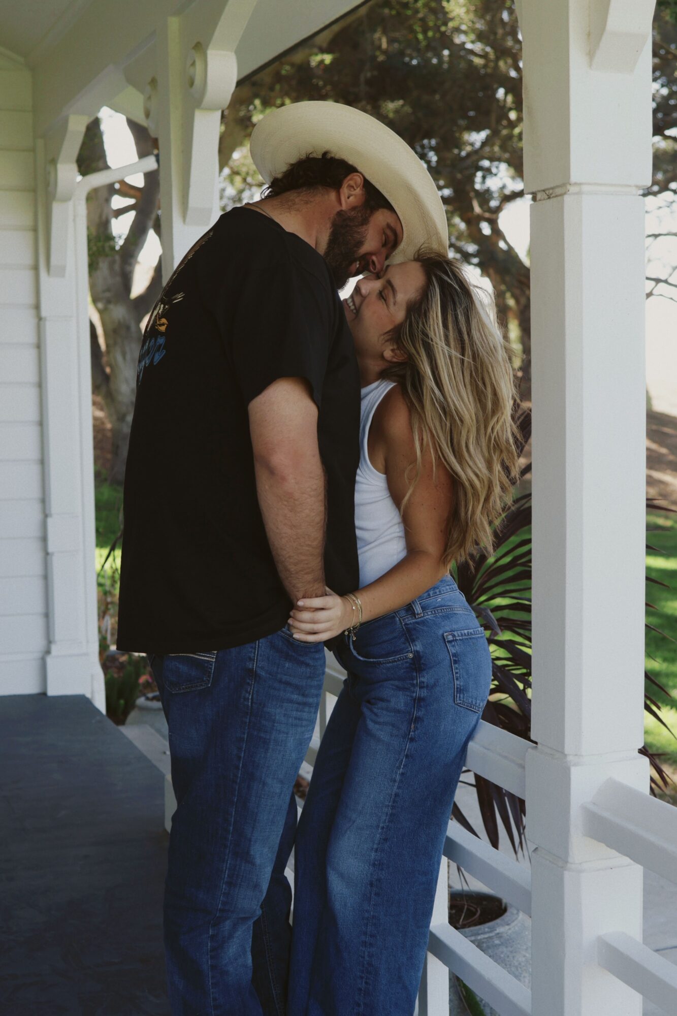 A man and woman are close together outdoors, with the woman leaning over the man who is wearing a cowboy hat, in a garden setting.