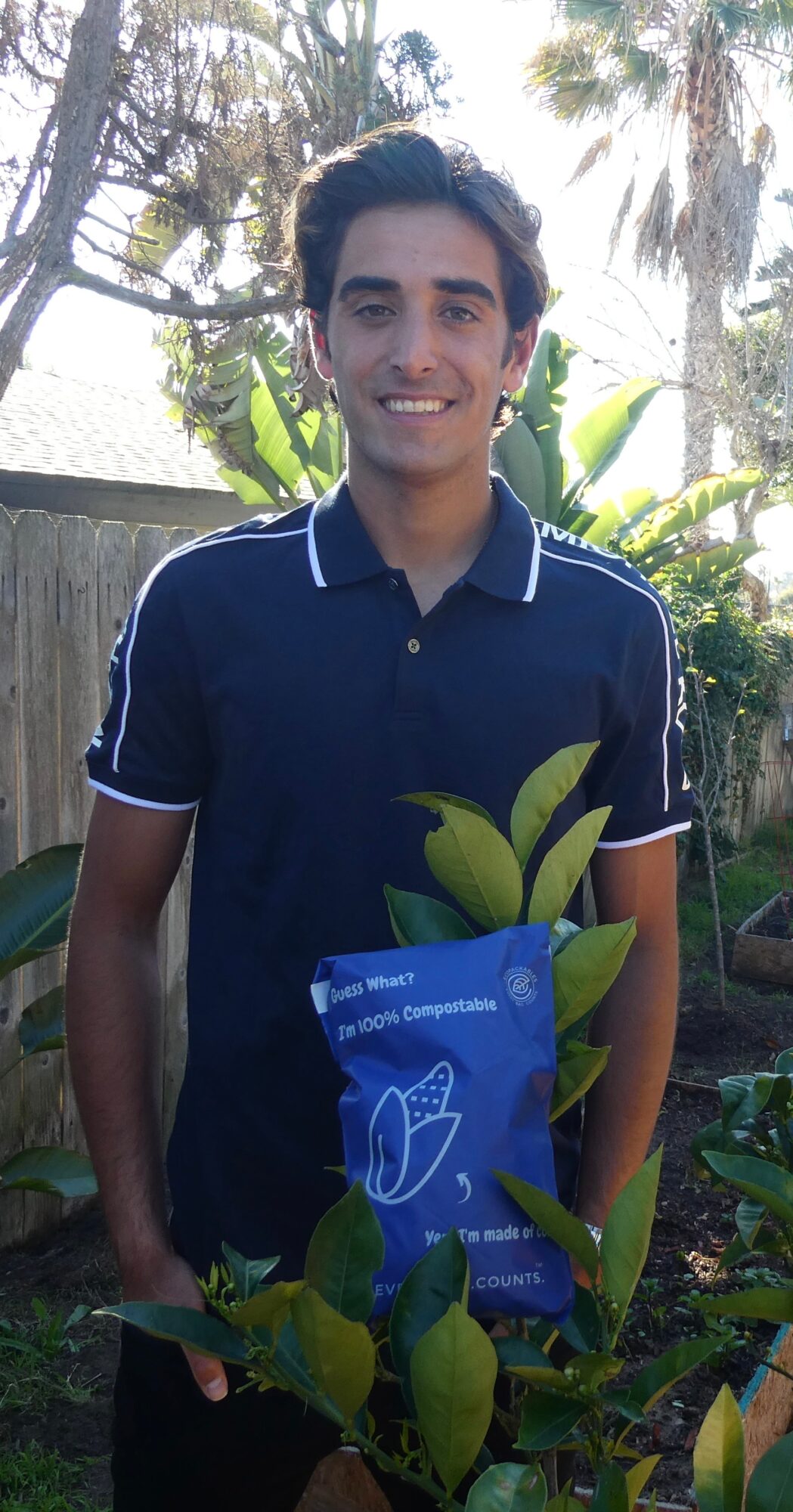 Young man standing outdoors with plants and trees, holding a blue book or folder, smiling at the camera.