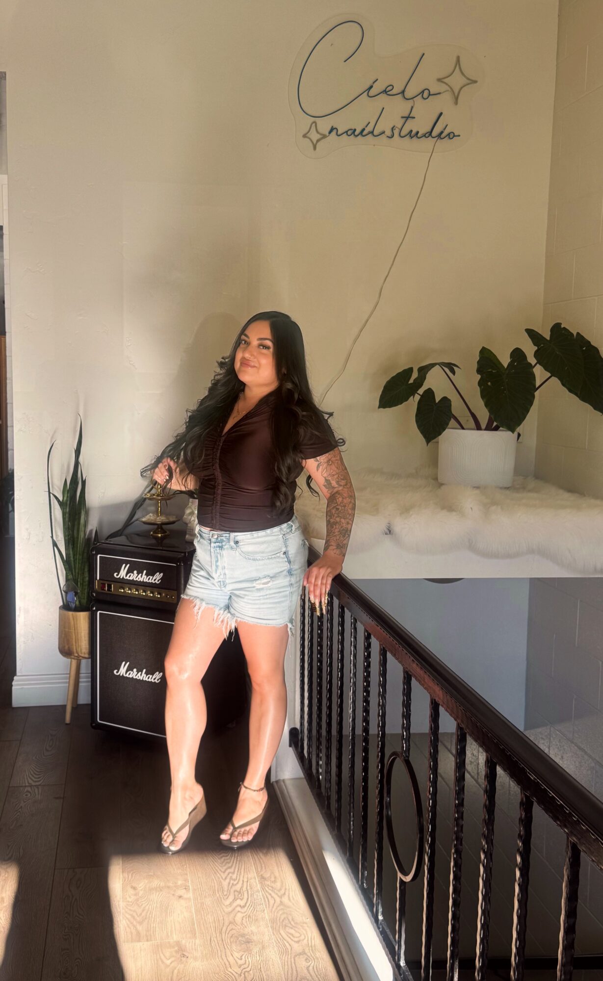 Woman with long dark hair wearing a brown top and denim shorts standing indoors near plants and Marshall amplifier, with handwritten wall art above.