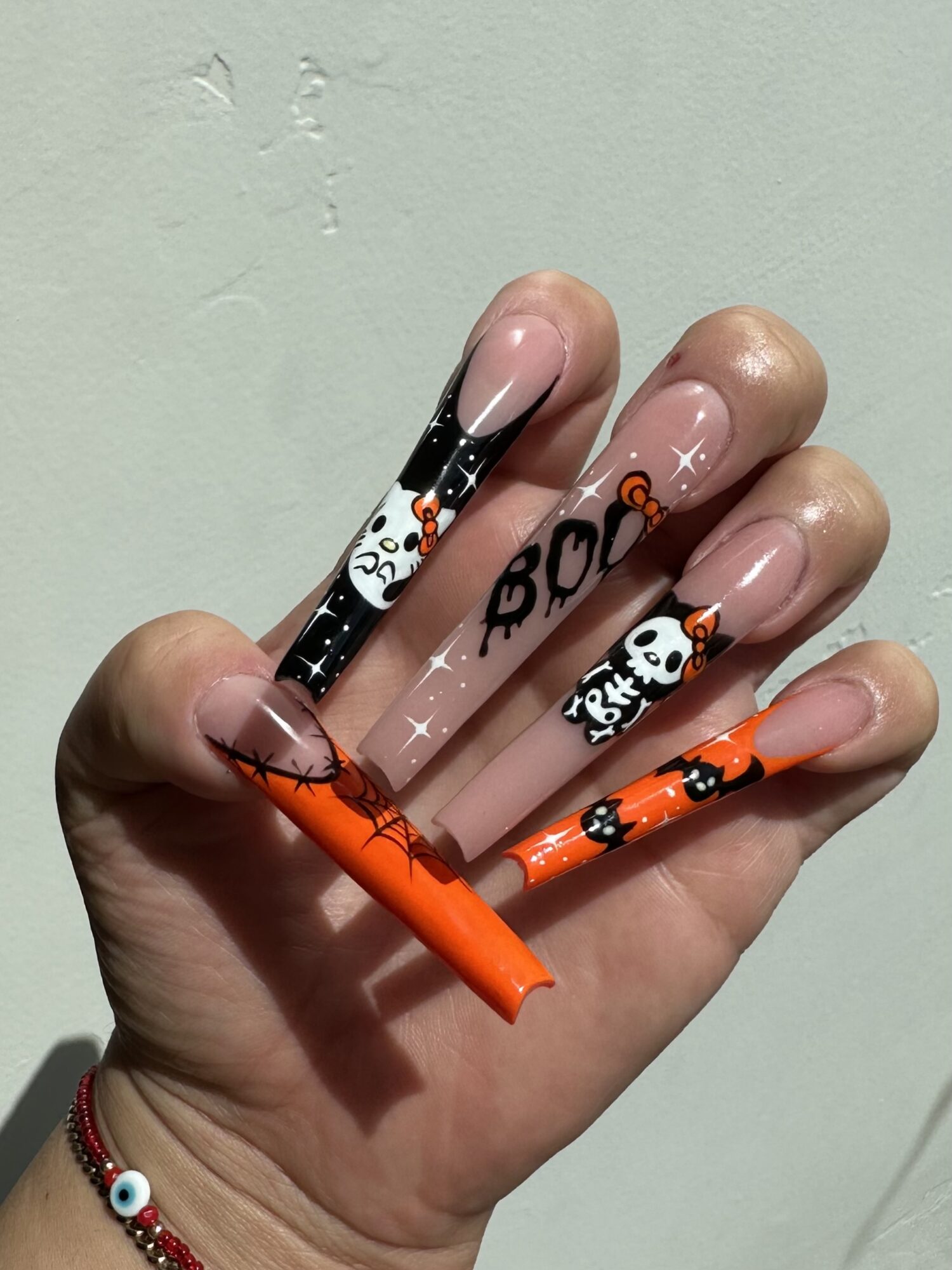 Hand with Halloween-themed long nails, decorated with ghosts, pumpkins, and spooky designs, against a plain wall background.