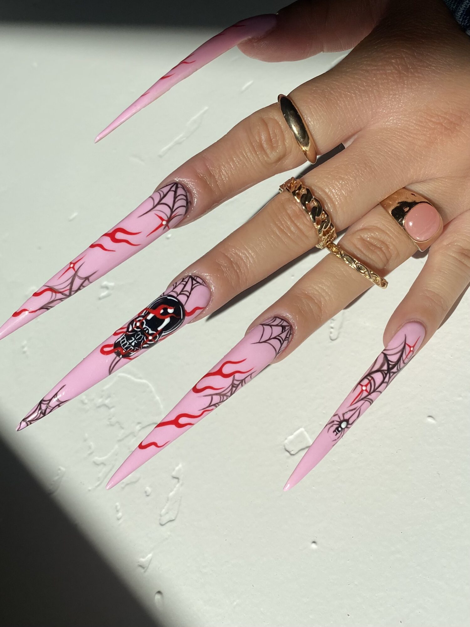 Hand with long, pointed pink nails decorated with black spiderweb and red flame designs, wearing multiple rings.