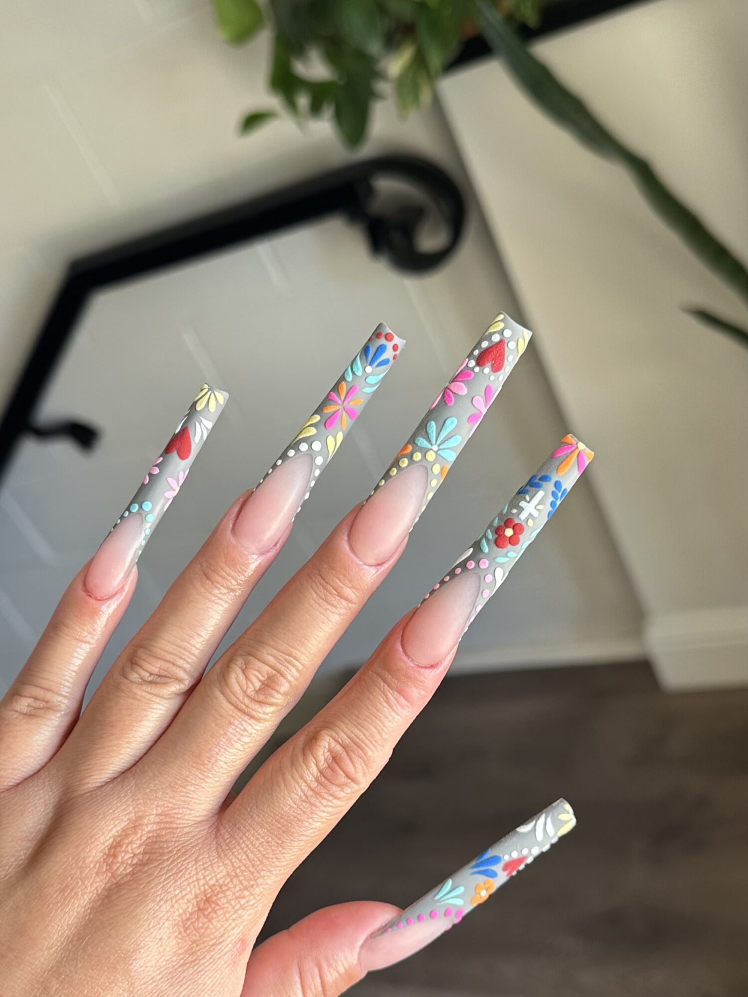 Hand with long, decorated nails featuring colorful floral and geometric patterns, against a blurred indoor background.