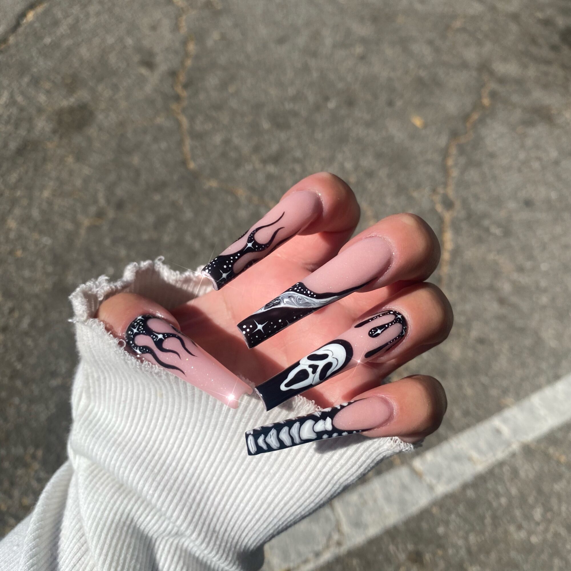 Hand with decorated nails in a white sleeve, outdoors on pavement, with a crack visible in the background.
