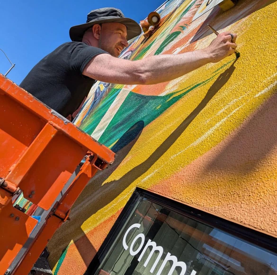 Man painting a colorful mural on a wall outdoors, wearing a hat and black shirt, with a bright blue sky background.