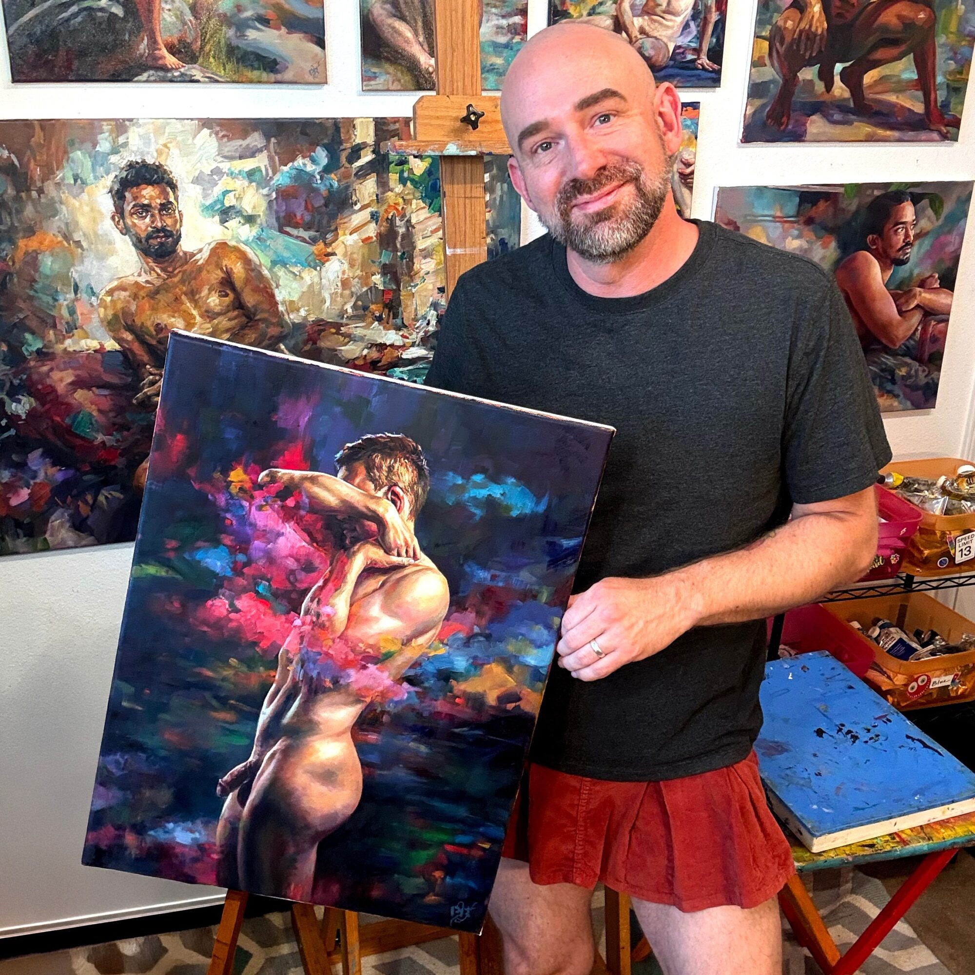 Man with a beard smiling, holding a colorful painting of a nude woman with clouds in the background, art studio setting.