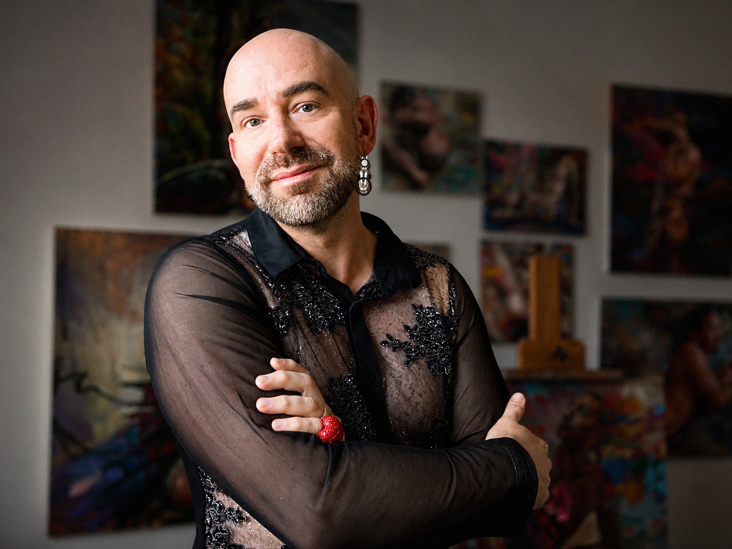 Bald man with beard wearing a black sheer top, standing with arms crossed in an art gallery.