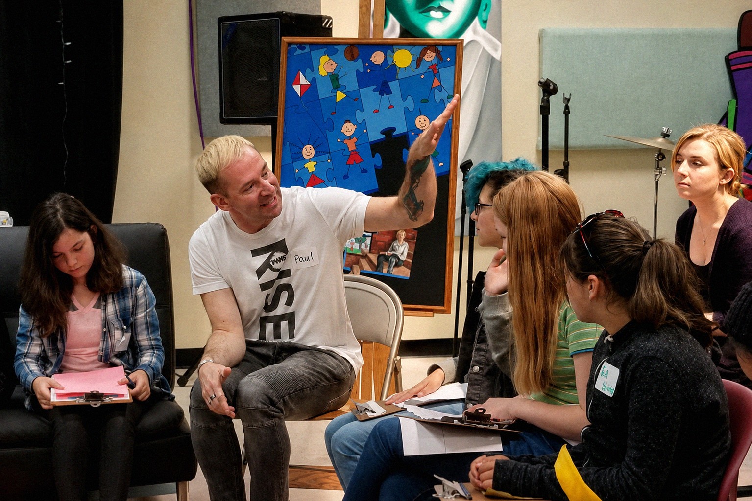 Man with blonde hair and tattoos gestures while speaking to a group of women in a room with a colorful poster and equipment.