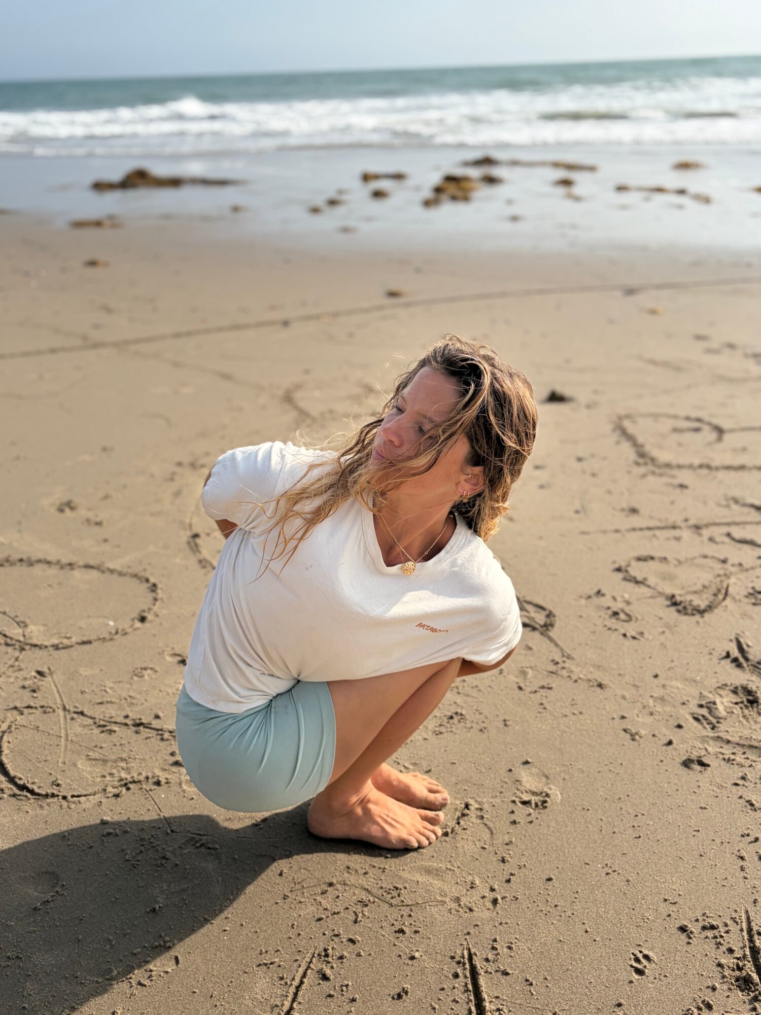 Woman crouching on sandy beach with ocean waves in background, writing in the sand, hair blowing in wind.