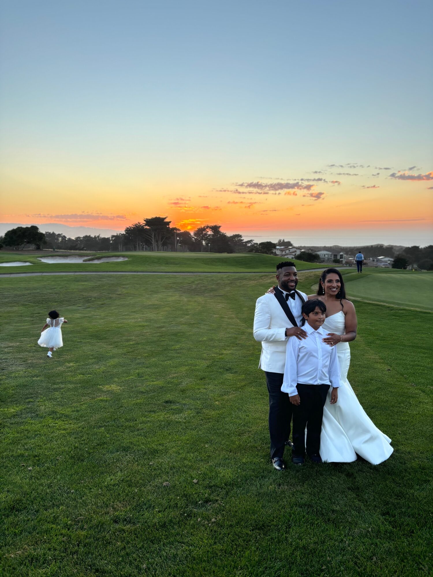 A family of four stands on a grassy field during sunset, with a child sitting on the grass nearby, sky with orange and blue hues.