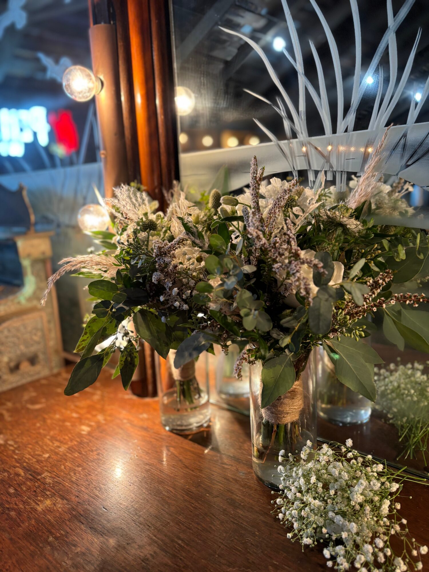 Two glass vases with white and purple flowers on a wooden surface near a window at night.