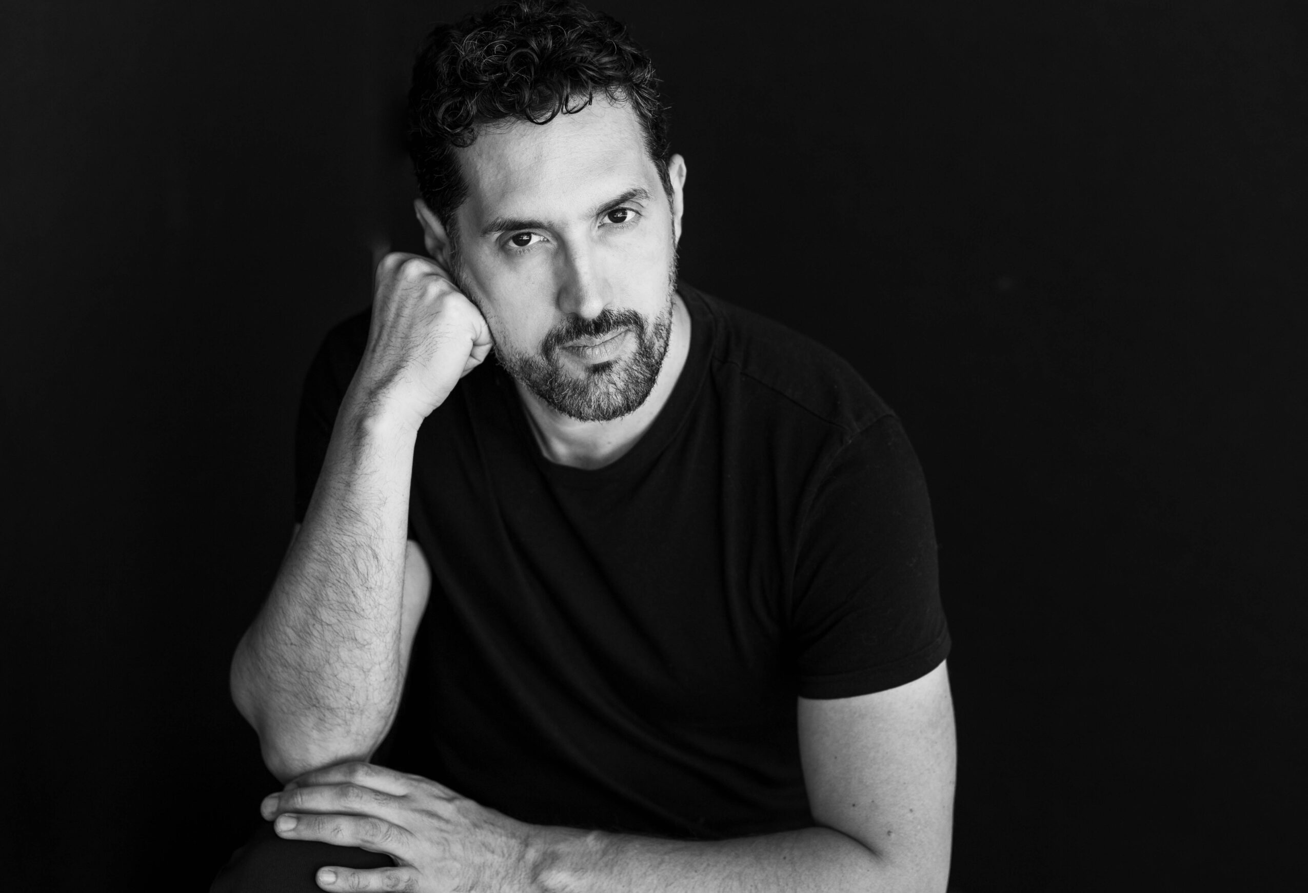 Man with curly hair and beard resting his head on his hand, wearing a black t-shirt, against a dark background.