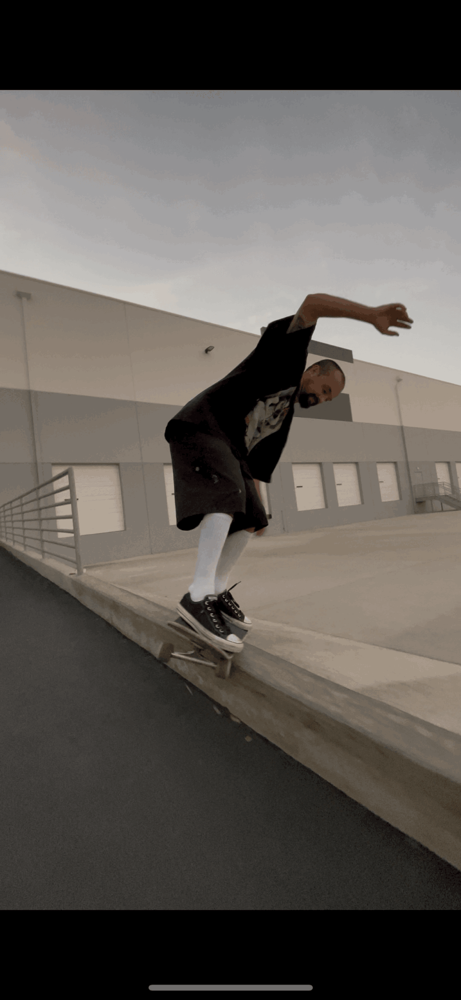 Person skateboarding on curb, leaning forward with arms outstretched, wearing black shirt, white pants, and black shoes.