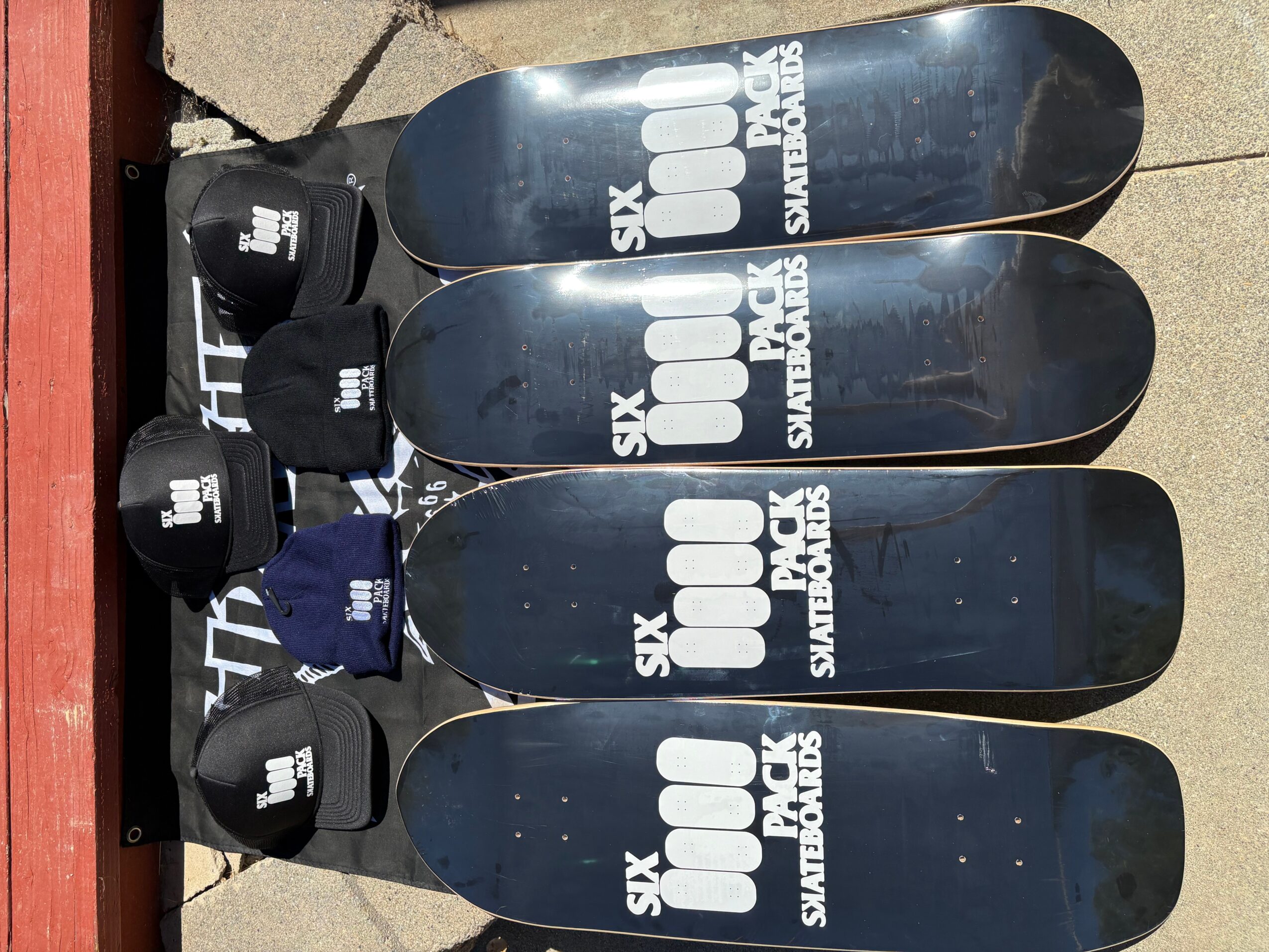 Four skateboards with black wheels and black grip tape, arranged on a concrete surface, with a red wooden edge nearby.