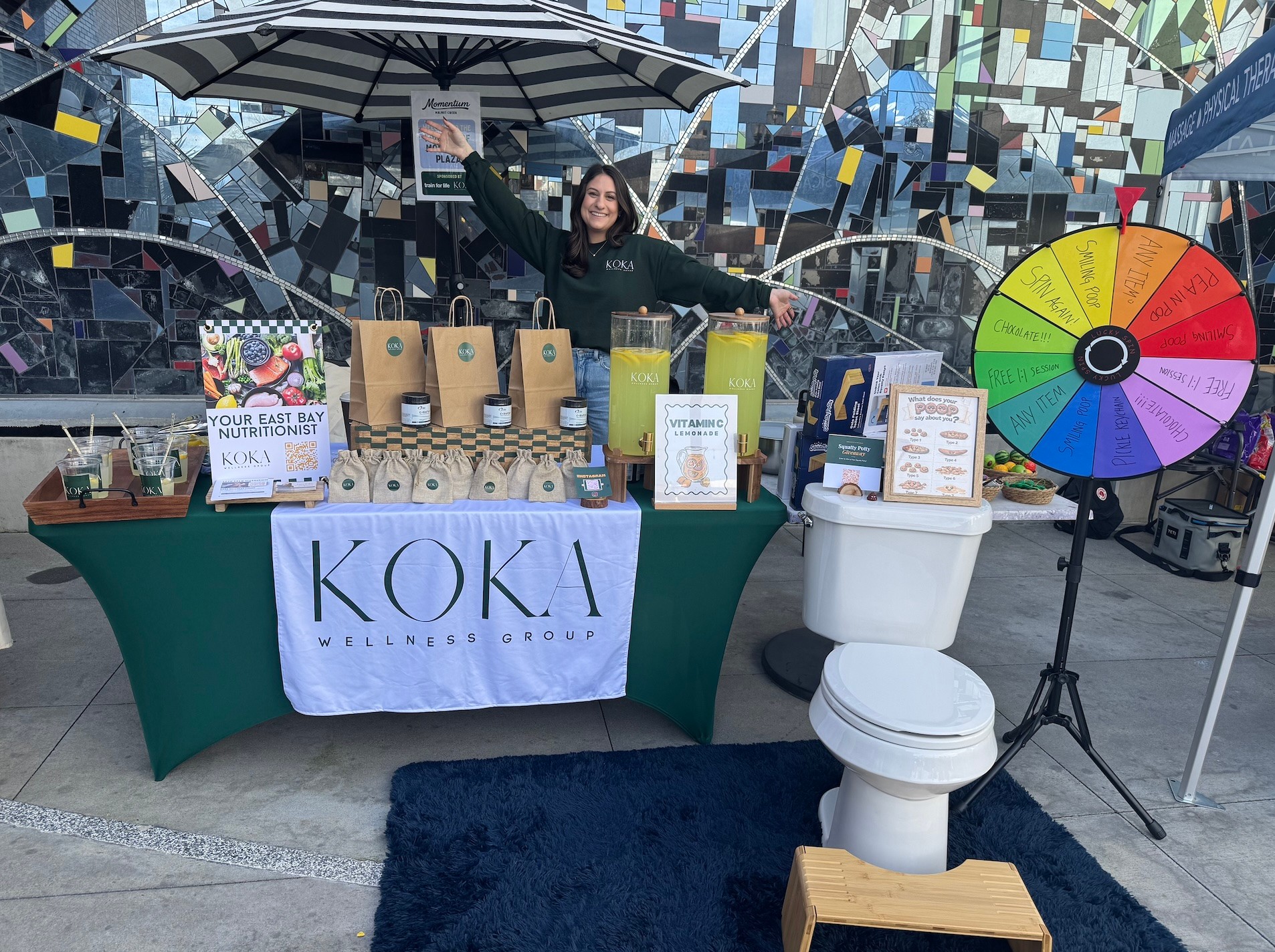 Woman standing behind a table with KOKA banner, promotional items, and a colorful spinning wheel outdoors.