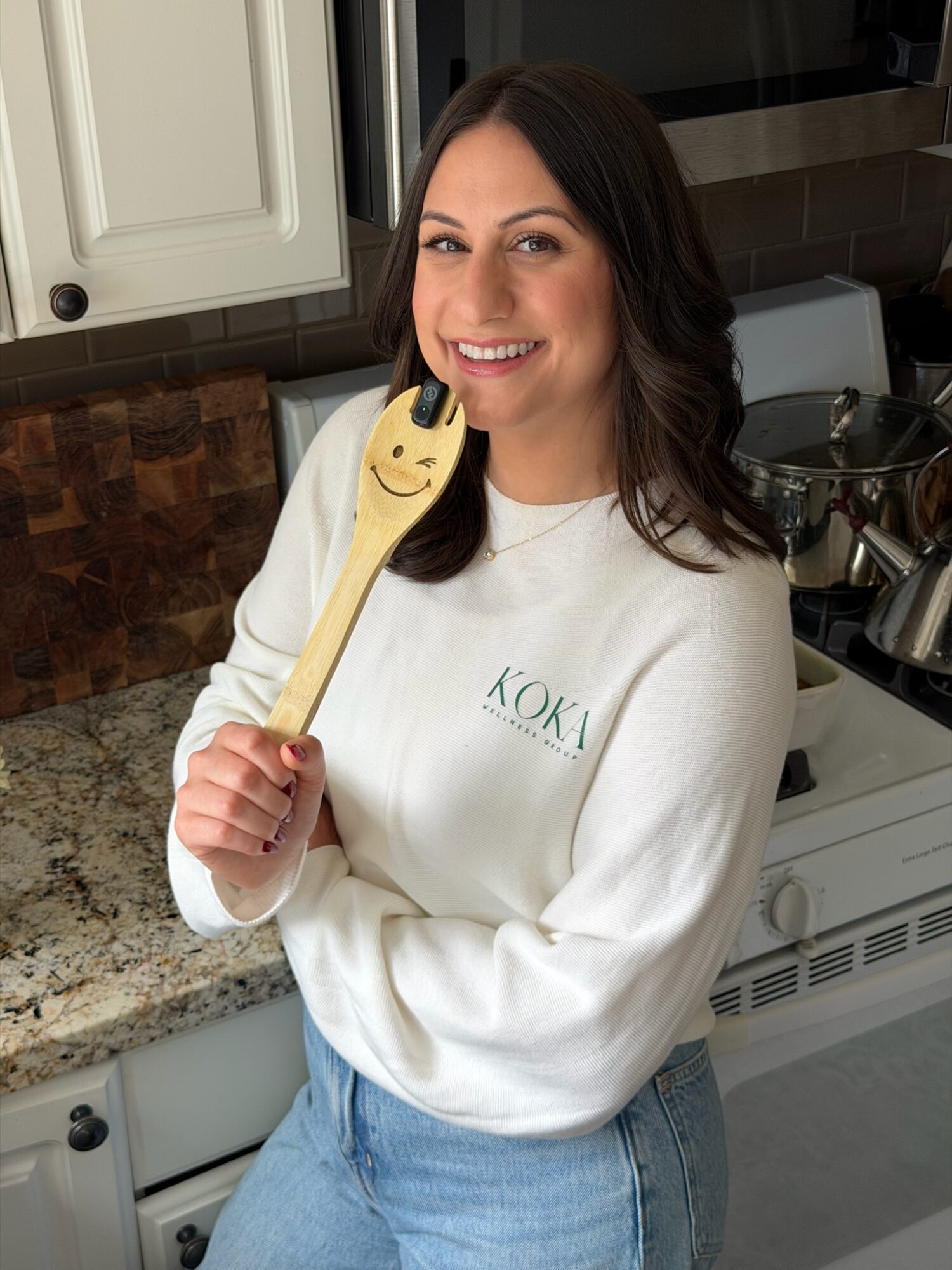 Woman with dark hair smiling in a kitchen, holding a wooden spoon with a smiley face, wearing a white sweatshirt and jeans.