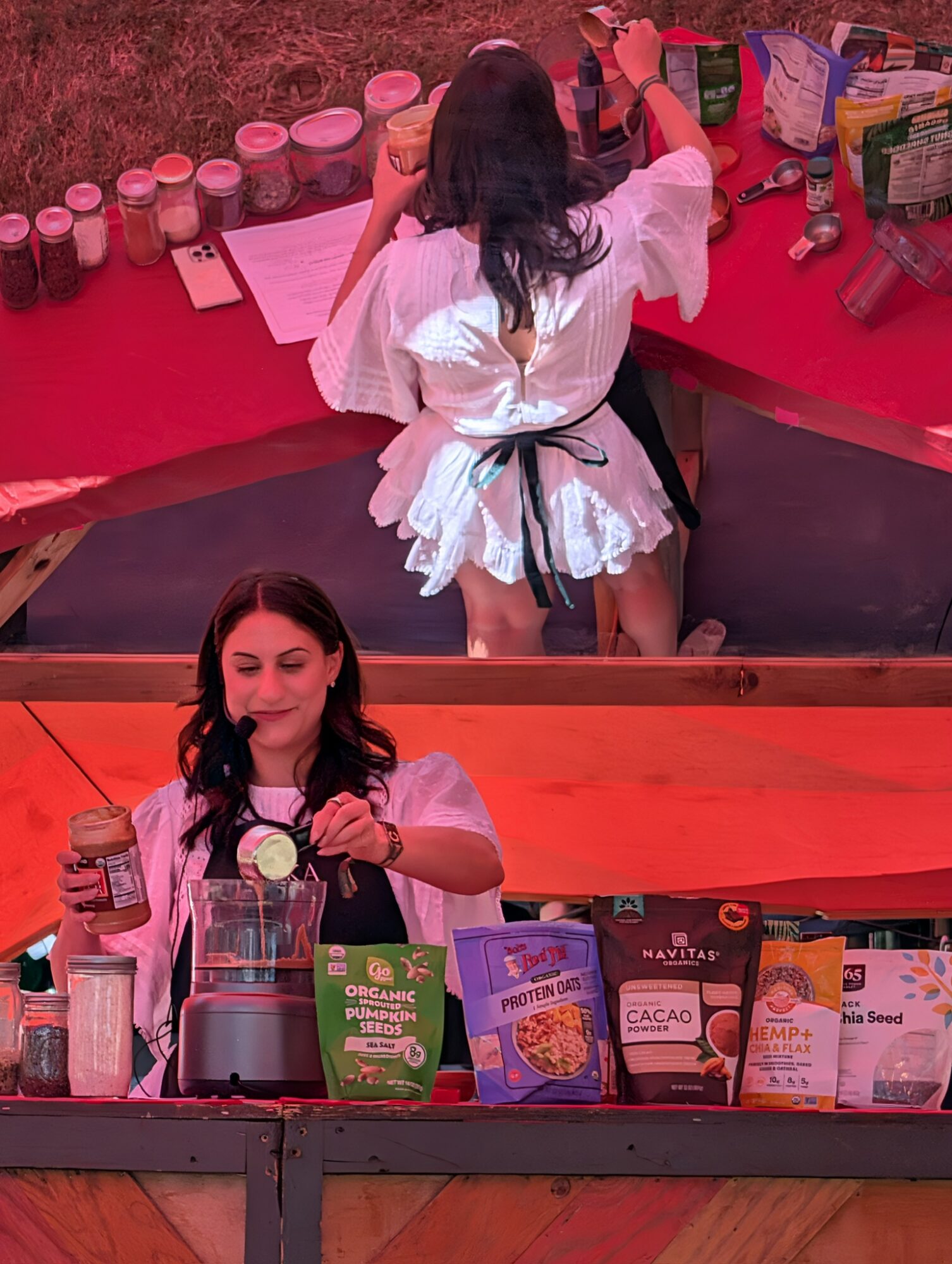 Two women preparing food at a table with various ingredients and packages, one blending ingredients, the other reaching for items.
