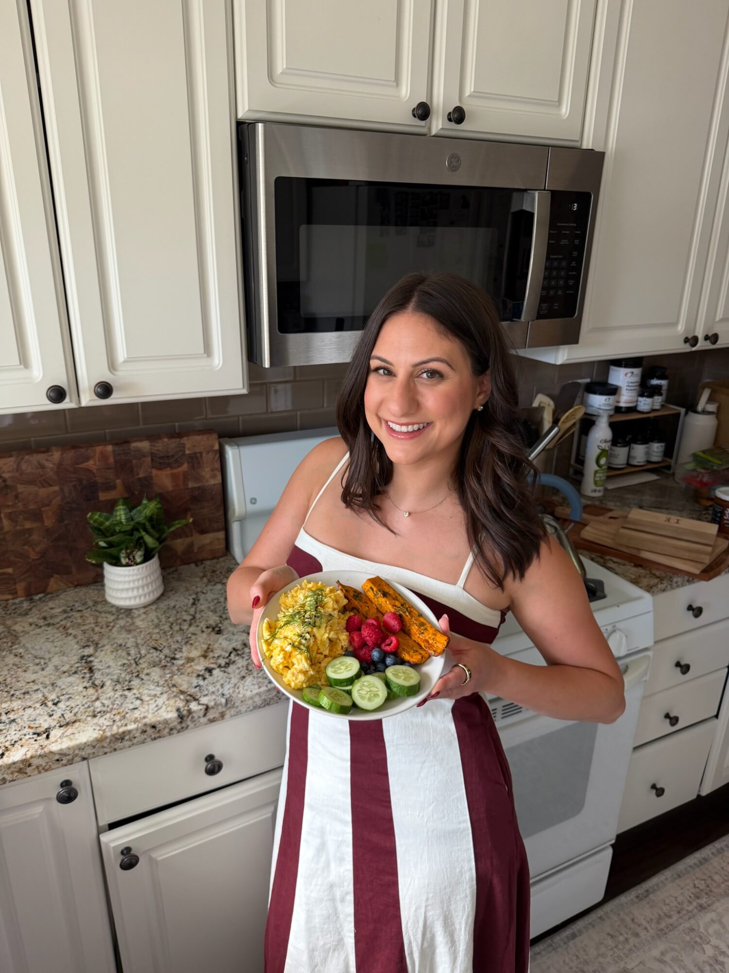 Woman in a striped apron holding a plate of food in a kitchen with white cabinets and a microwave.