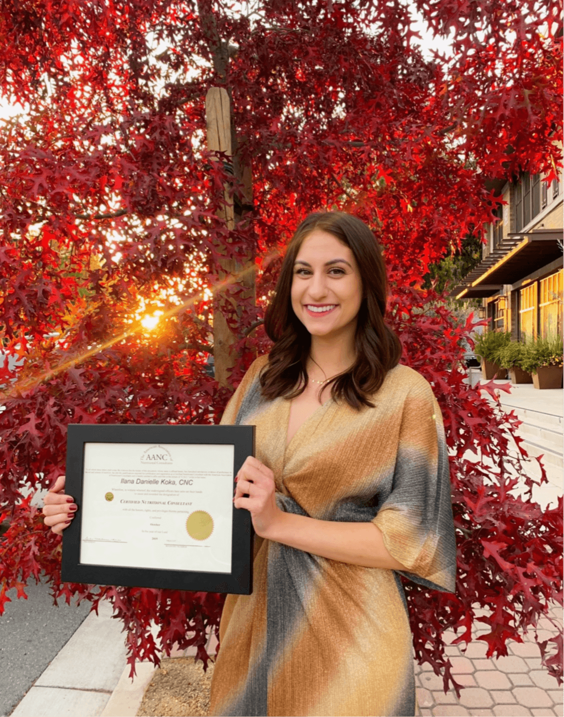 Young woman holding a diploma in front of red autumn leaves, smiling outdoors during sunset.