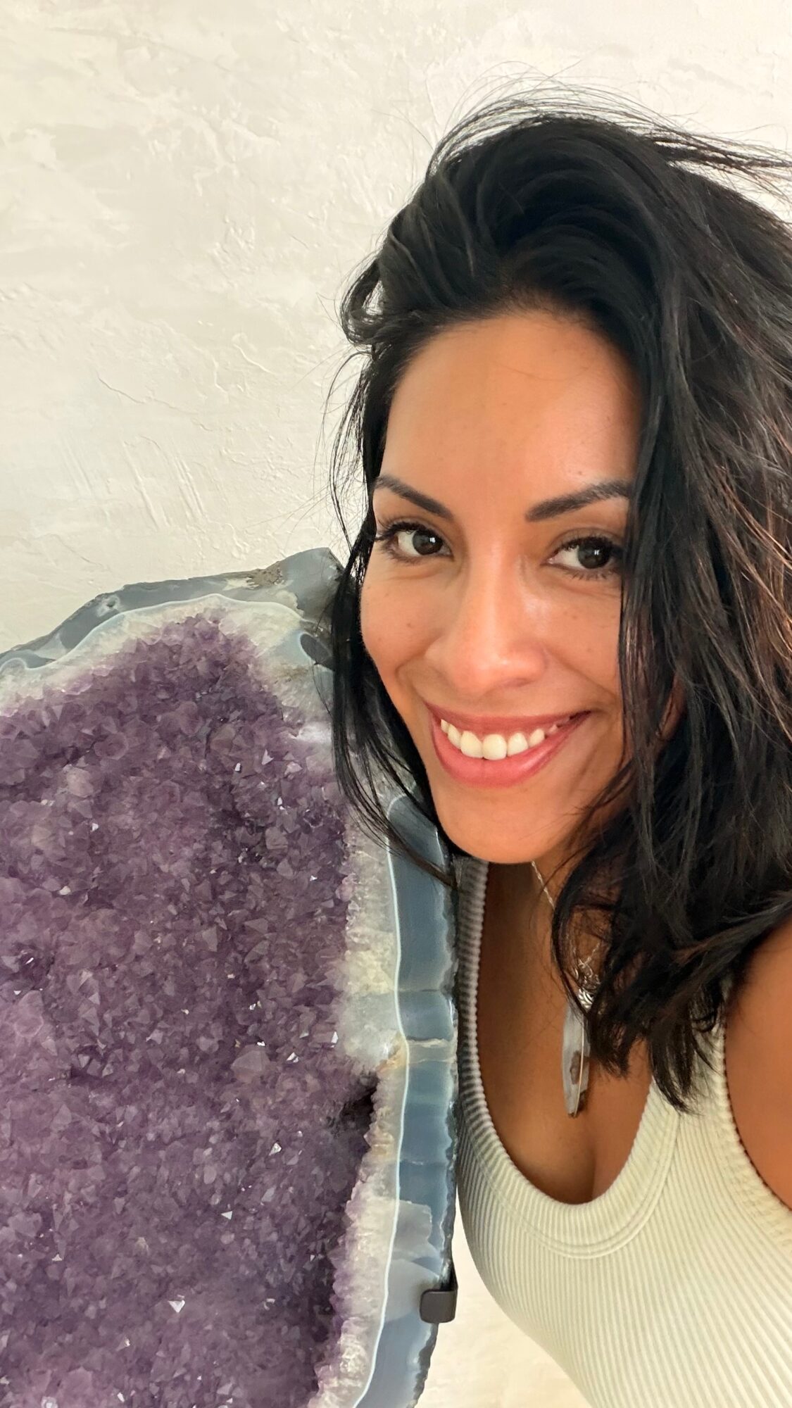 Smiling woman with dark hair and white top, holding a purple crystal cluster, against a light background.