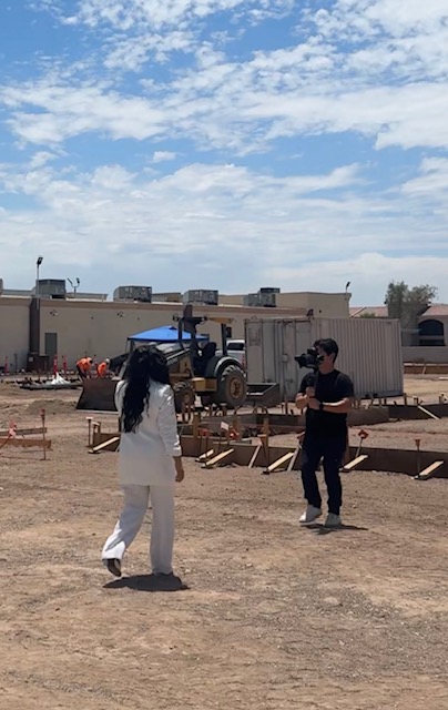 Two people standing outdoors on a construction site with a tractor and buildings in the background under a partly cloudy sky.