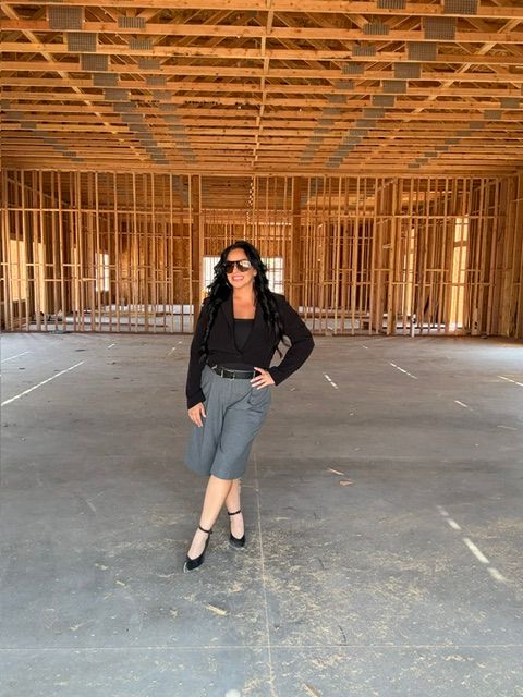 Woman standing in an indoor space with wooden wall panels, wearing a black jacket, gray skirt, and black shoes.