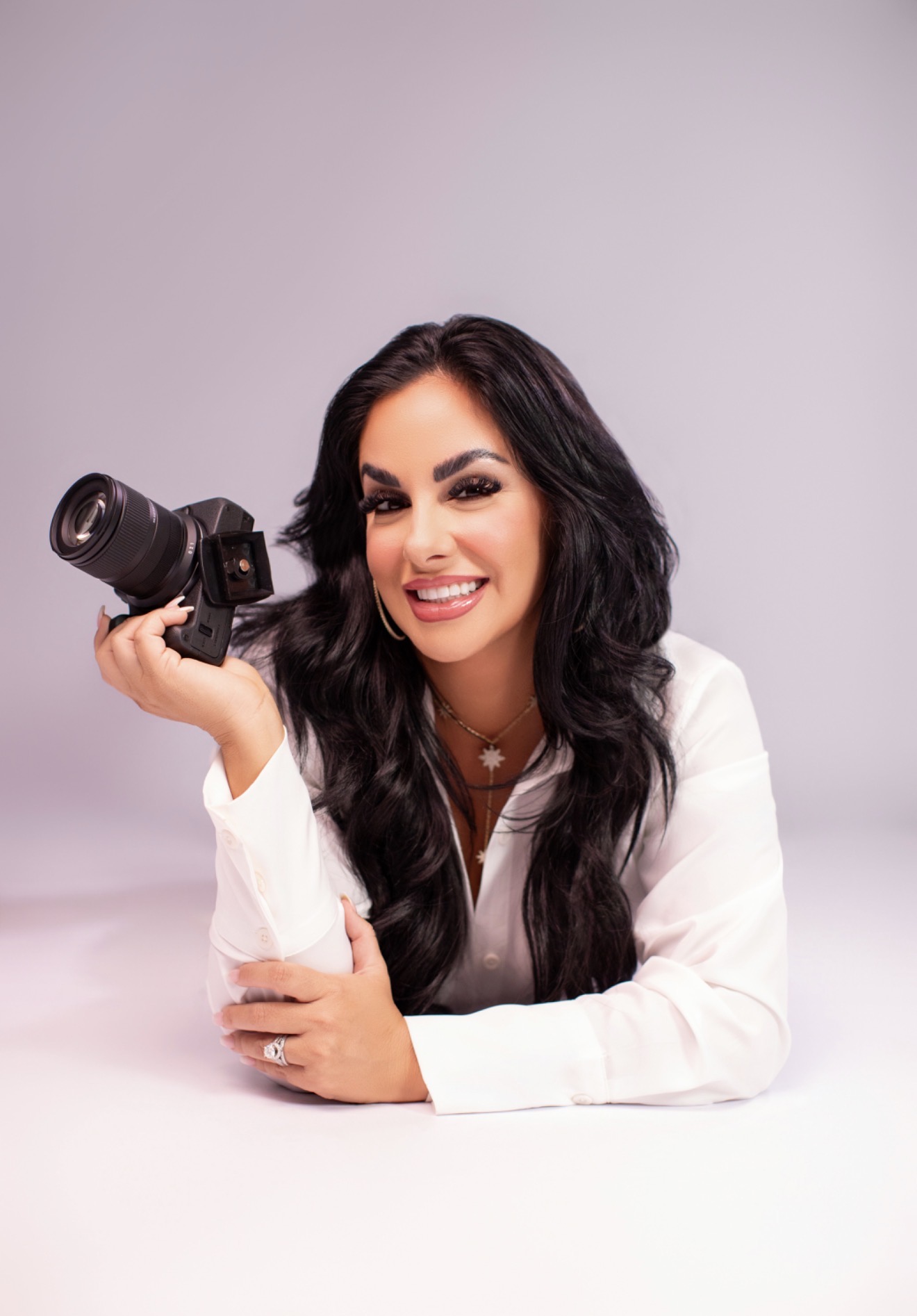 Woman with long dark hair smiling, holding a camera, wearing a white shirt, against a plain light background.