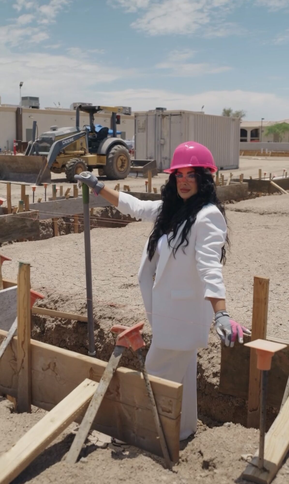 Woman in white coat and pink helmet using a tool at construction site with equipment and buildings in background.