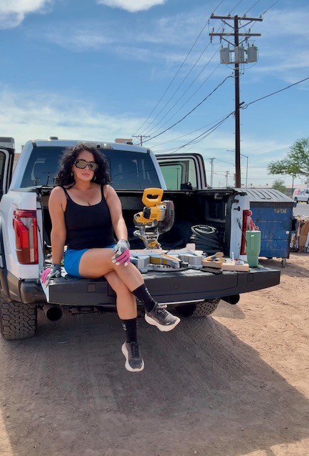 Woman sitting on truck tailgate with tools and equipment, outdoors on dirt ground, blue sky, power lines, and trees in background.