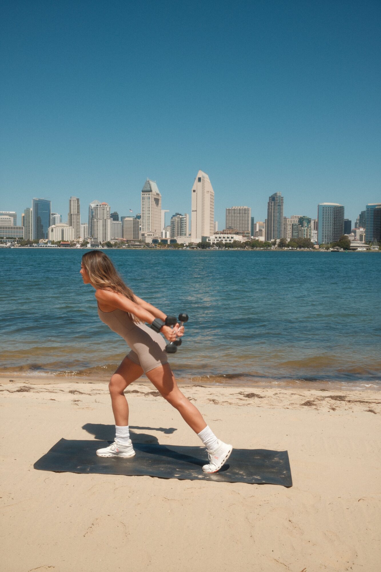 Person with long hair on a surfboard at the beach, city skyline in the background, clear blue sky.
