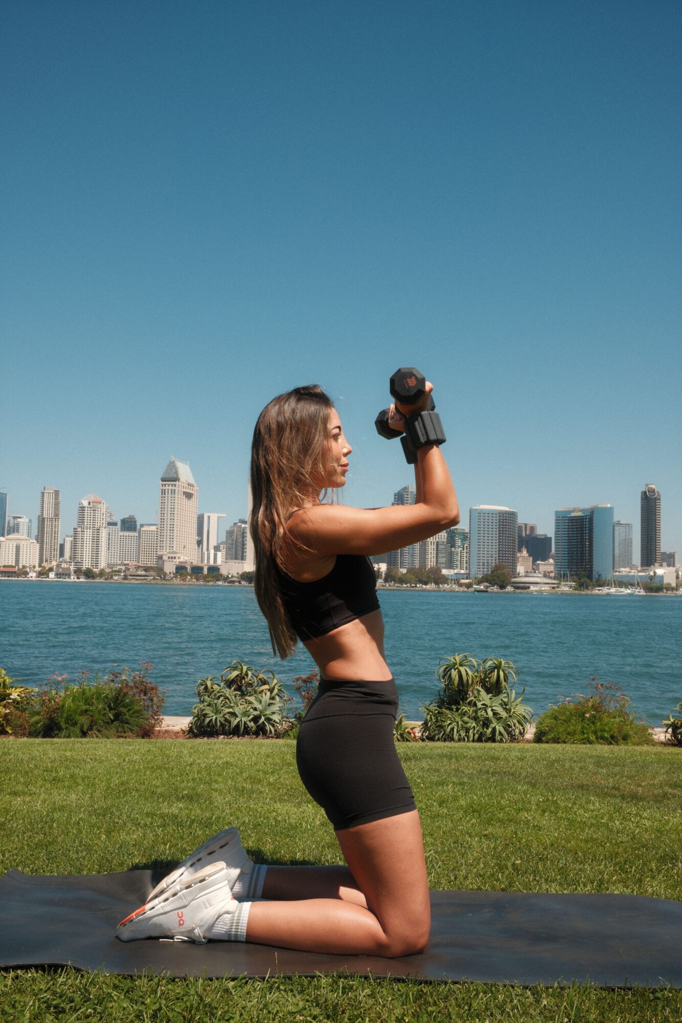 Woman doing push-ups outdoors near water with city skyline in background, wearing black sports bra and shorts.