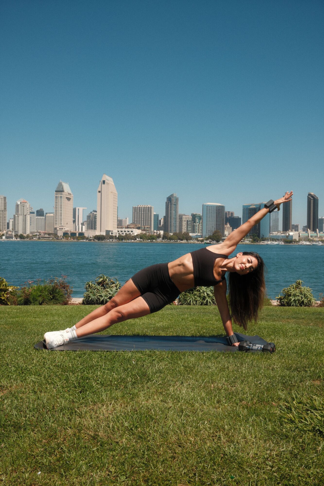 Woman practicing yoga outdoors on a mat with city skyline and water in background, sunny day.